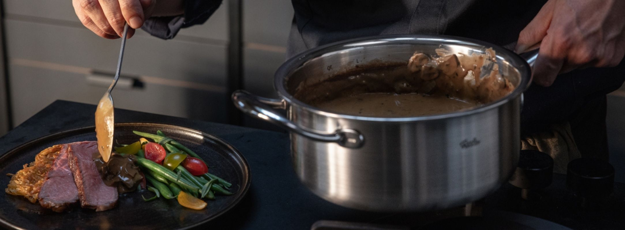 Ein Heimkoch serviert in einer modernen Küche Sauce aus einem Fissler-Edelstahltopf auf einen Teller mit geschnittenem Fleisch und Gemüse.