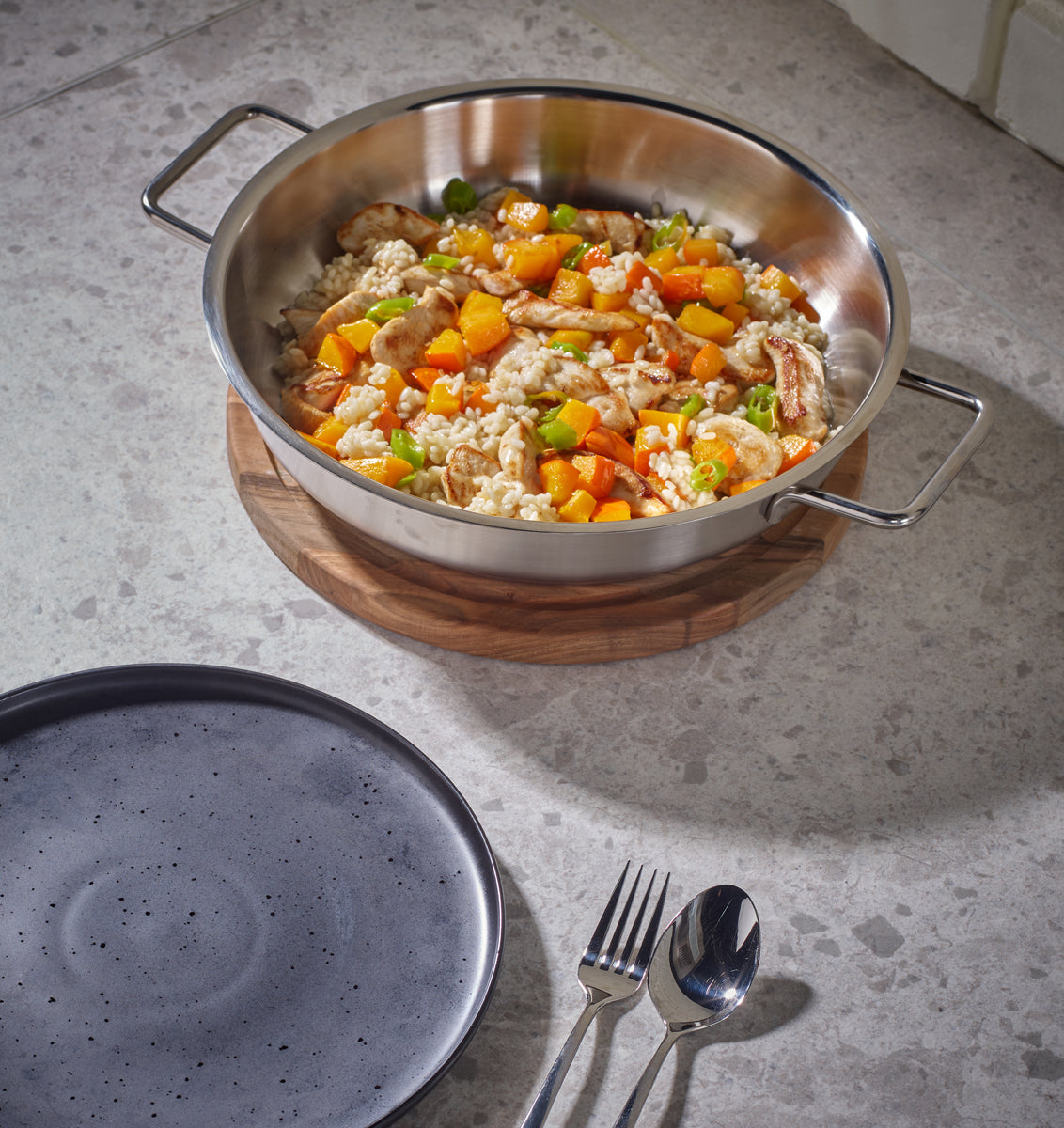 Fissler Pure serving pan, made in Germany from recycled stainless steel, filled with rice, vegetables, chicken; plate and cutlery next to it.