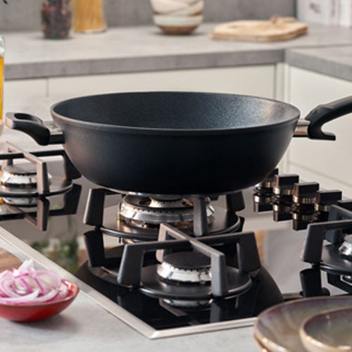 Der Fissler Adamant® Wok mit kratzfester Beschichtung auf einem Gasherd, der deutsche Präzision und Langlebigkeit demonstriert. Rote Zwiebeln in der Nähe.