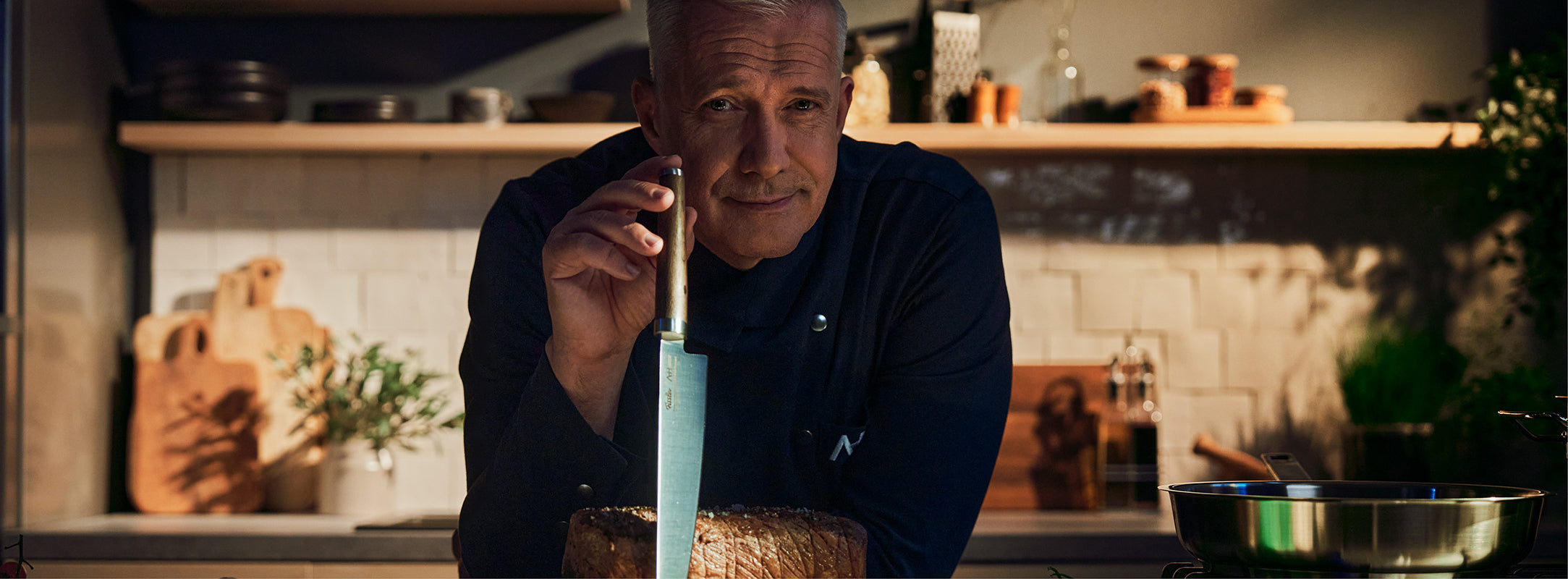Ein Koch in dunkler Uniform steht in einer Küche und präsentiert ein Fissler-Messer und ein Brett mit Brot, dahinter ist hochwertiges Kochgeschirr zu sehen.