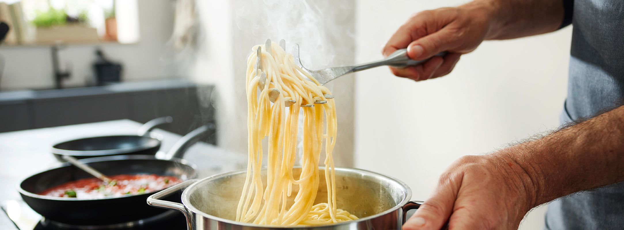 Eine Person hebt dampfende Spaghetti mit einer Zange aus einem Fissler-Edelstahltopf; im Hintergrund steht ein Fissler-Topf mit Tomatensauce.