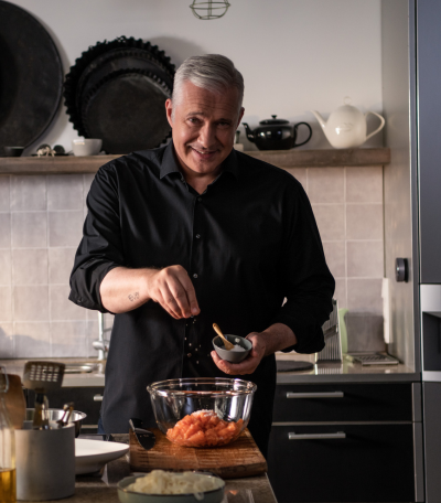 Ein Mann in einem schwarzen Hemd lächelt, während er in einer modernen Küche Hackfleisch würzt, während hinter ihm Fissler-Kochgeschirr und -Utensilien fein säuberlich angeordnet sind.