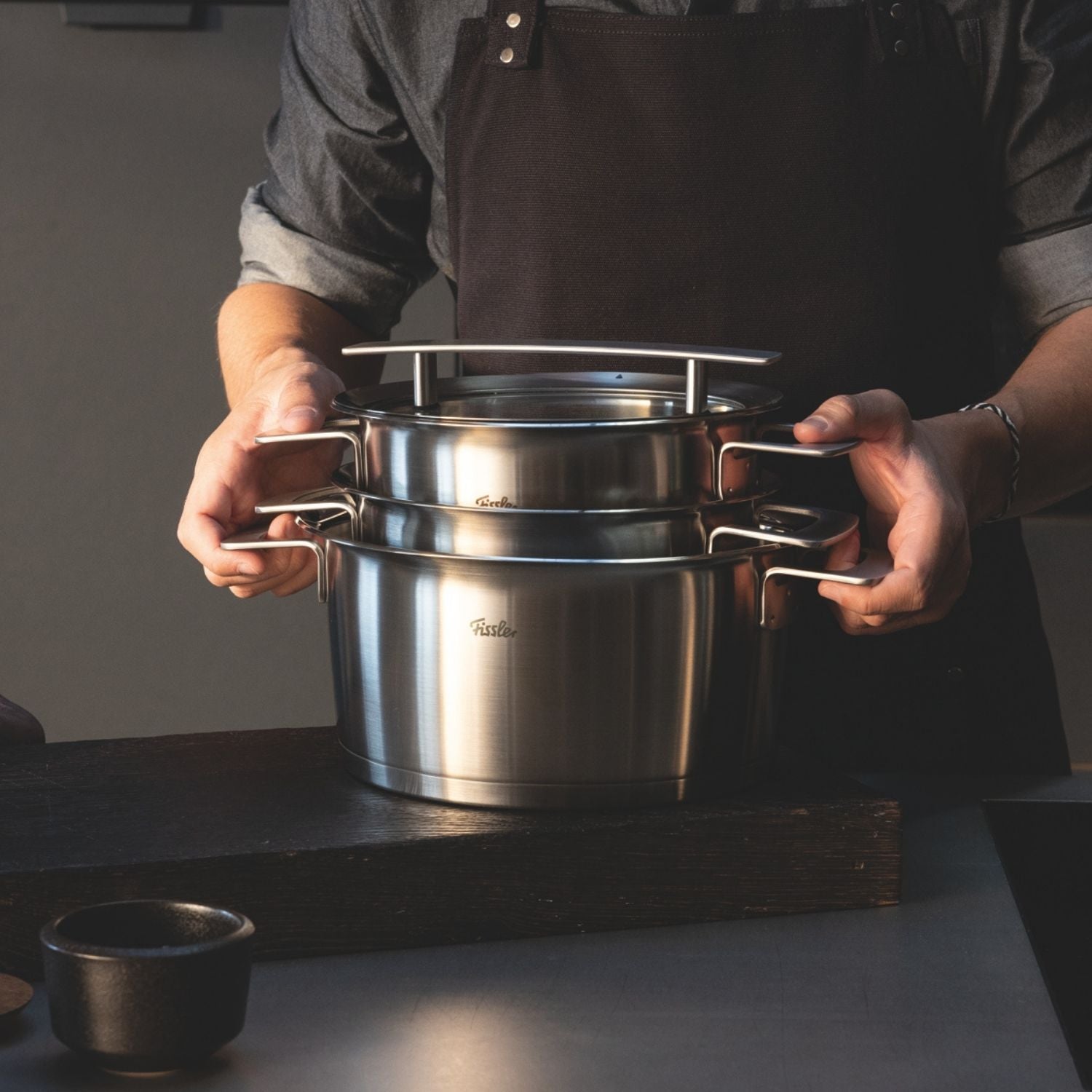 Eine Person in dunkler Schürze hält zwei übereinander gestapelte Fissler-Edelstahltöpfe mit Deckeln und präsentiert deutsche Qualität auf einer schlichten Küchenzeile.