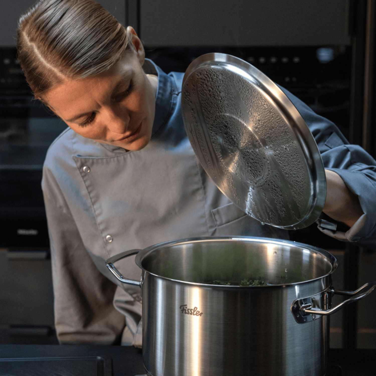 Ein Koch in Grau hebt den Deckel eines Fissler-Edelstahltopfes an, während Dampf aufsteigt, und präsentiert damit deutsche Innovation und zeitloses Design.