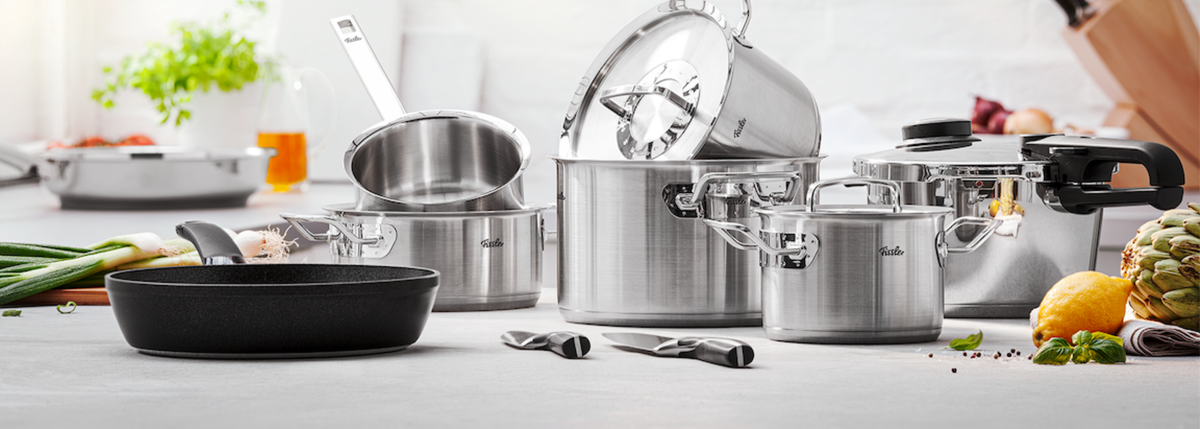 Töpfe, Pfannen und Messer aus rostfreiem Stahl von Fissler auf einer hellen Küchentheke, dahinter frisches Gemüse, Kräuter und Gewürze.