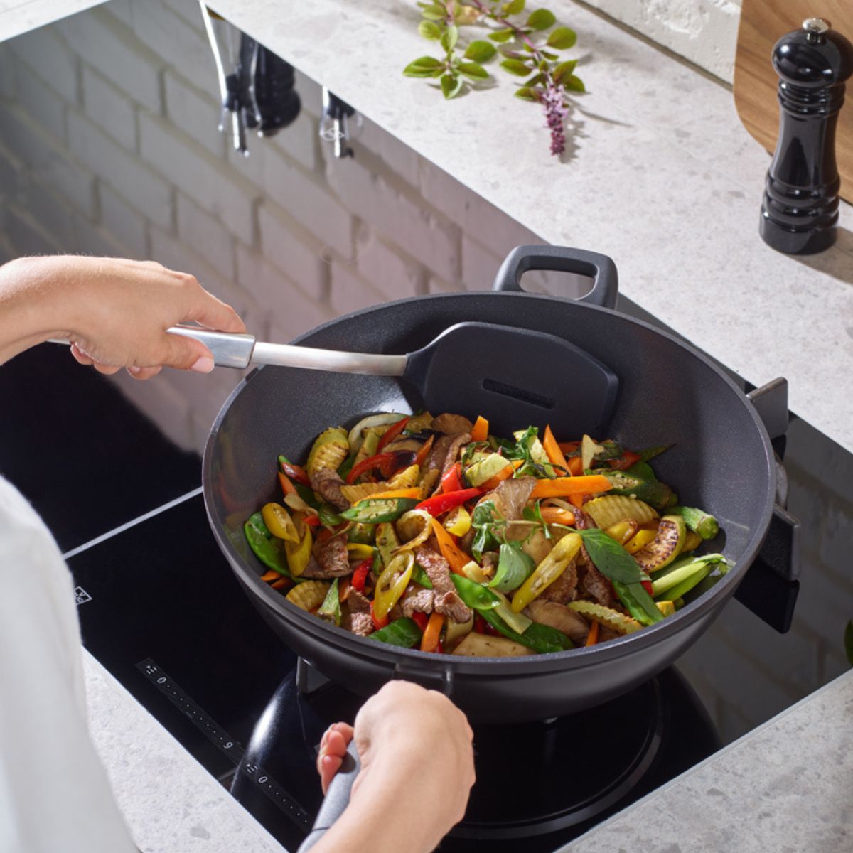 Eine Person brät Gemüse und Fleisch in einem Fissler-Wok auf einem Elektroherd; Kräuter und Pfeffermühle liegen auf dem Tresen daneben.