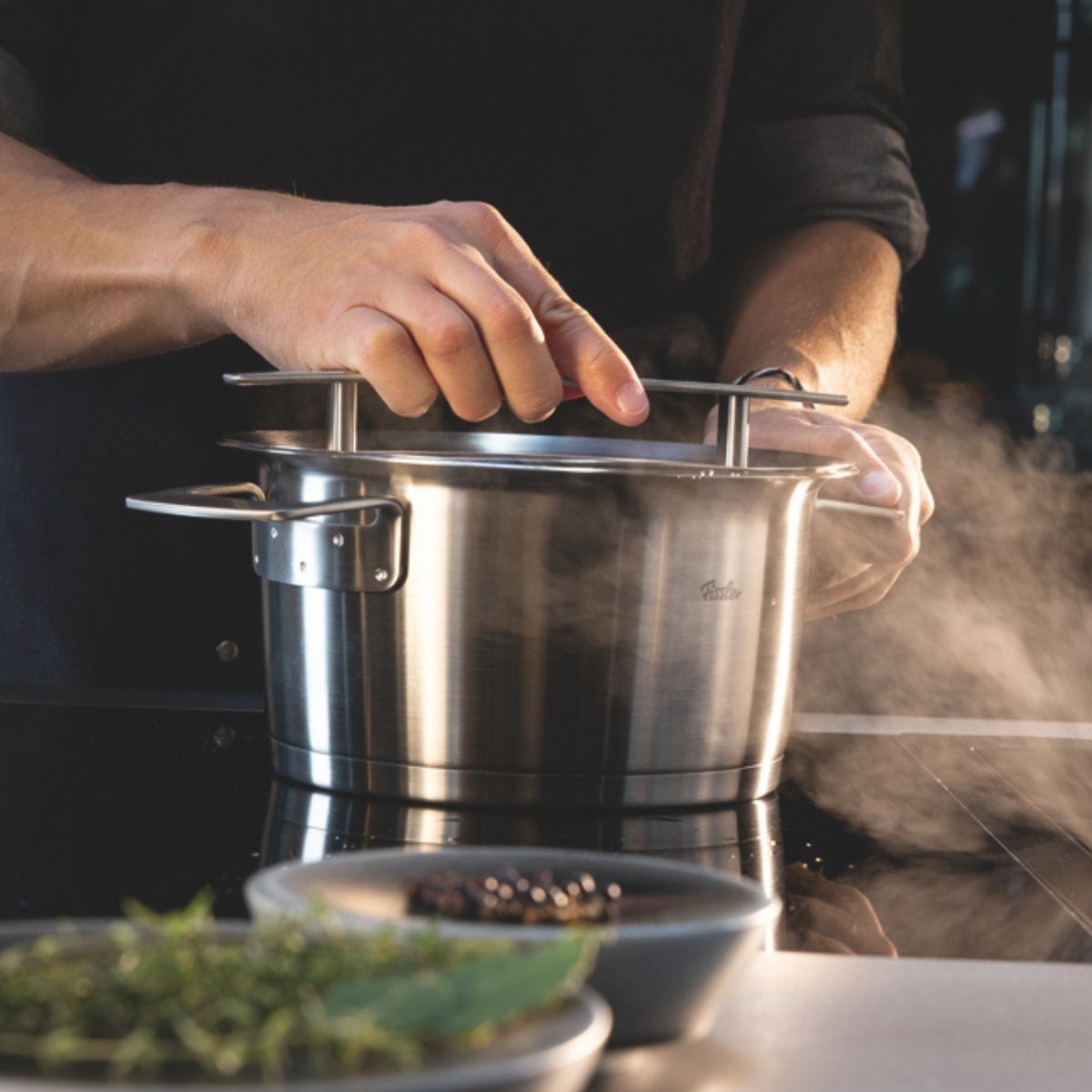 Eine Person hebt den Deckel von einem Fissler-Edelstahltopf auf dem Herd, in dem frische Kräuter und Zutaten für die Gourmetküche bereitstehen.