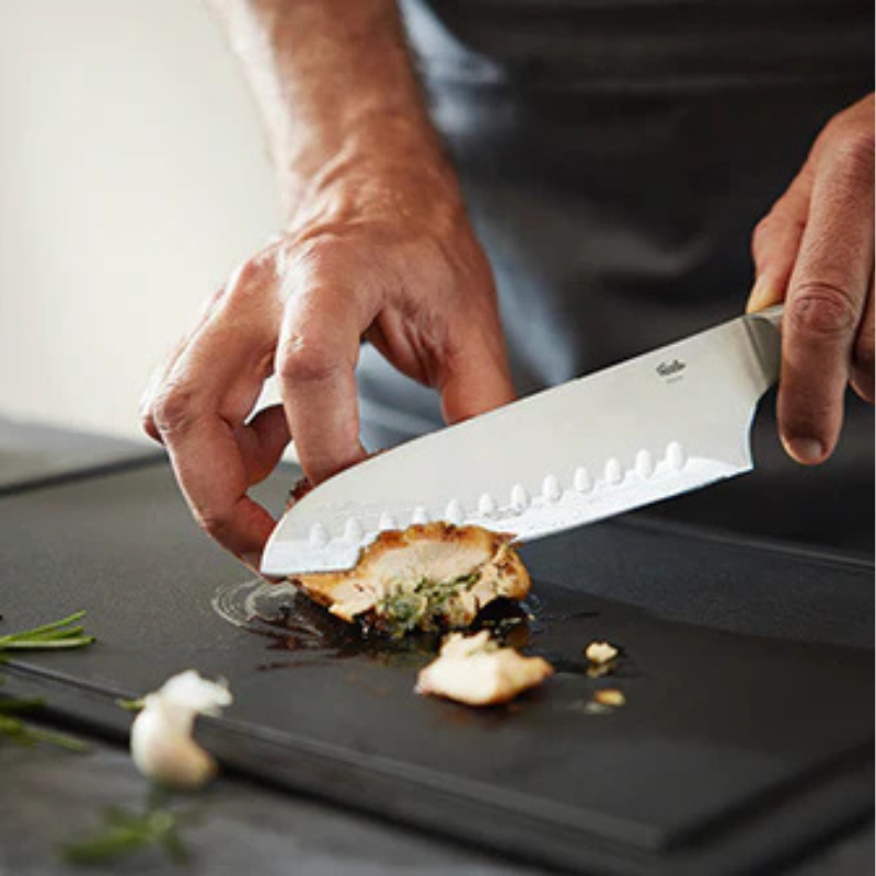 Eine Person schneidet mit einem Fissler-Messer eine mit Kräutern gefüllte Hähnchenbrust auf einem schwarzen Brett präzise in Scheiben; Rosmarin und Knoblauch liegen daneben.