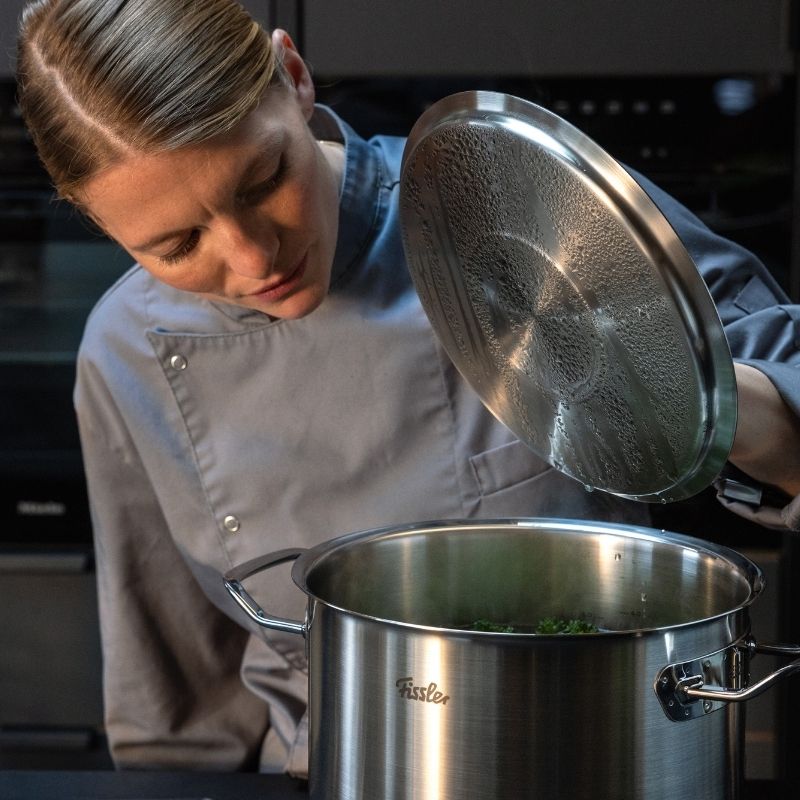 Ein Koch hebt den Deckel eines Fissler-Edelstahltopfs auf der Theke an, der Dampf ist sichtbar - ein Musterbeispiel für deutsche Präzisionskochgeschirre.