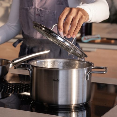 A person lifts the lid of a Fissler stainless steel pot, developed in Germany, as steam rises; another Fissler pot in the background.