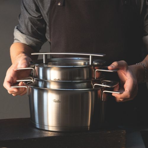 A person in a dark apron holds two stacked Fissler stainless steel pots with sturdy handles and lids, presented on a smooth dark surface.