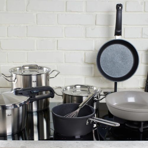 Stainless steel pots, pans, and a whisk by Fissler on a stovetop in a modern kitchen with a white brick backsplash, showcasing German quality.