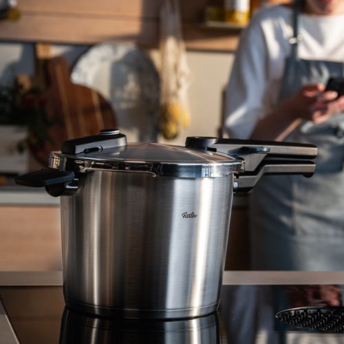 A Fissler stainless steel pressure cooker sits on a stovetop in an elegant kitchen; a blurred chef in an apron stands behind it holding a phone.