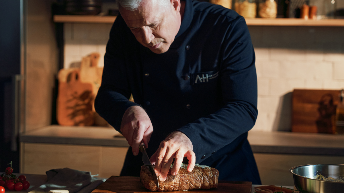 Ein Koch in dunkler Uniform schneidet fachmännisch Brot auf einem Holzbrett, Fissler-Kochgeschirr und Utensilien sind in der warm beleuchteten Küche zu sehen.