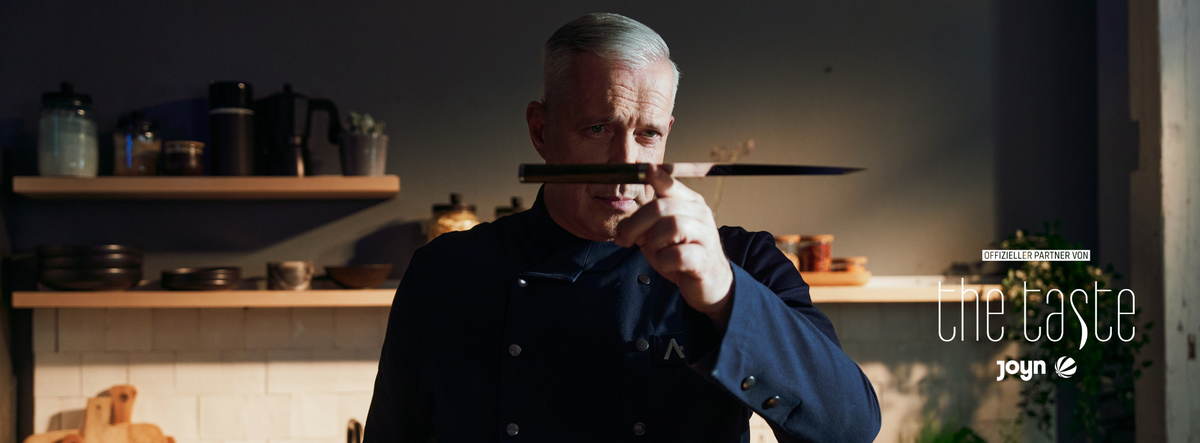 Ein Koch in dunkler Uniform prüft ein Fissler-Küchenmesser auf Augenhöhe in einer modernen Küche, der Text "the taste" und "Joyn" ist sichtbar.