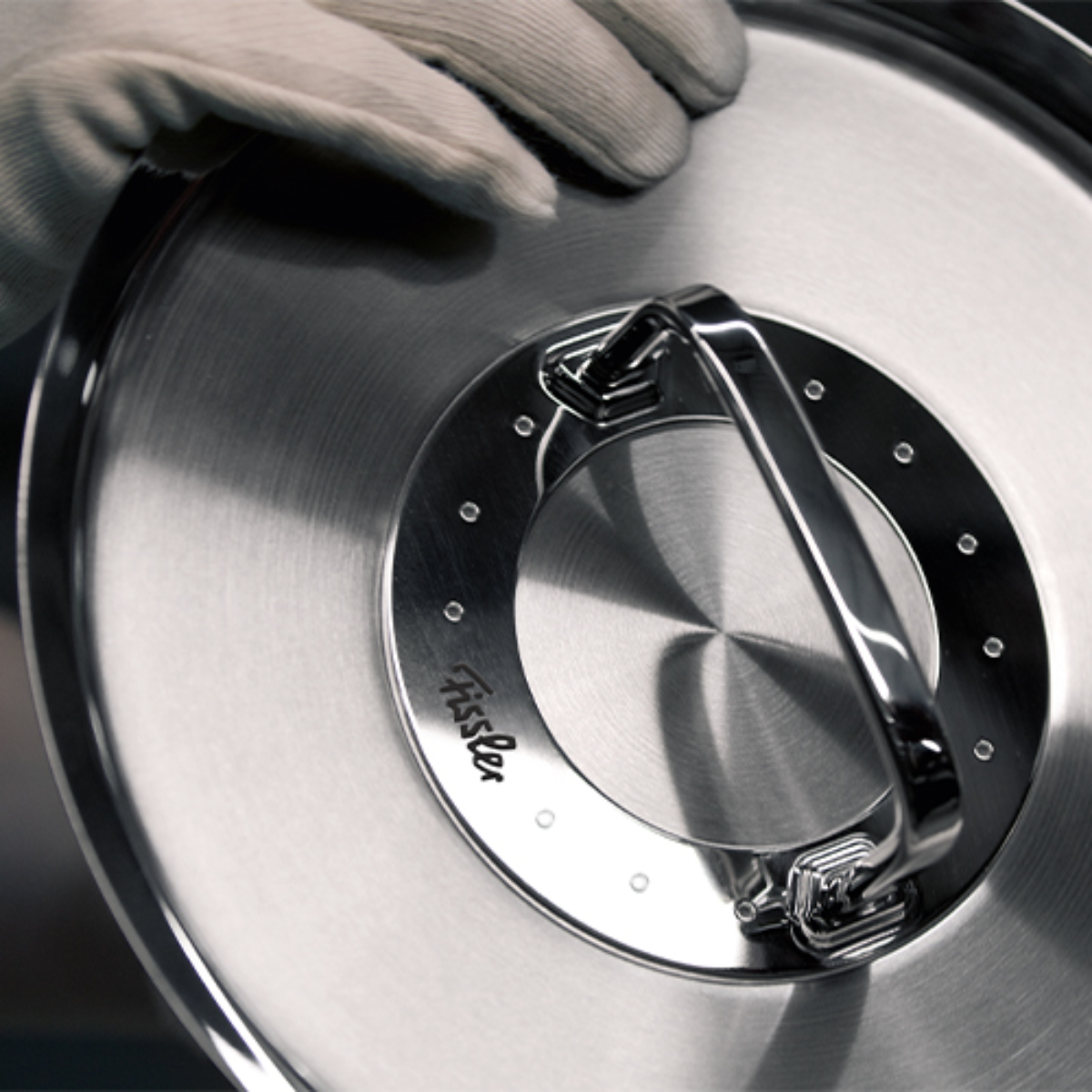 Eine behandschuhte Hand hält einen hochwertigen Fissler-Edelstahldeckel mit einem großen Griff und eingraviertem Logo - deutsche Präzisionsarbeit.