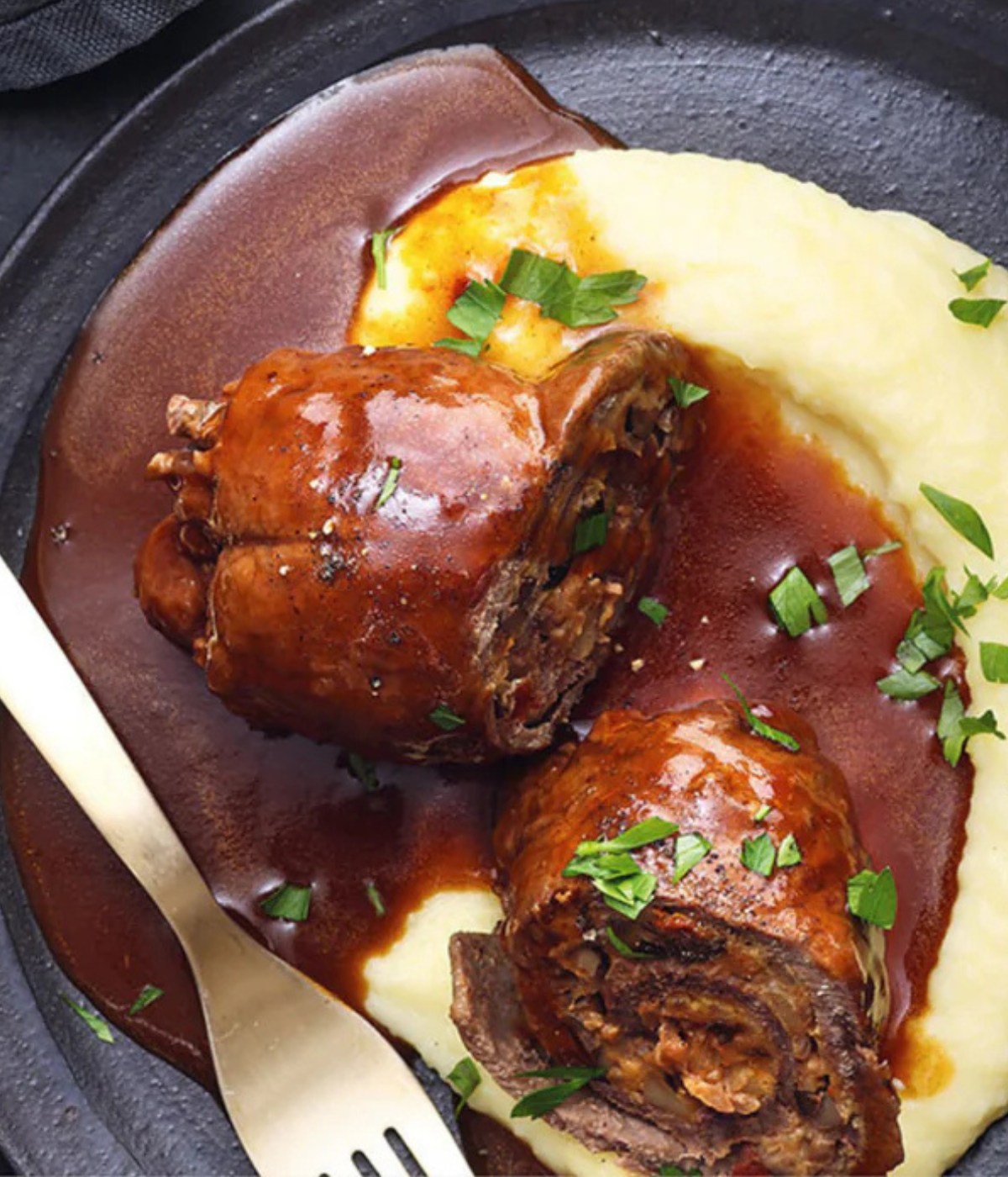 Tender braised beef roulade in brown sauce over mashed potatoes, herb garnish; served on a dark plate - prepared with Fissler precision.