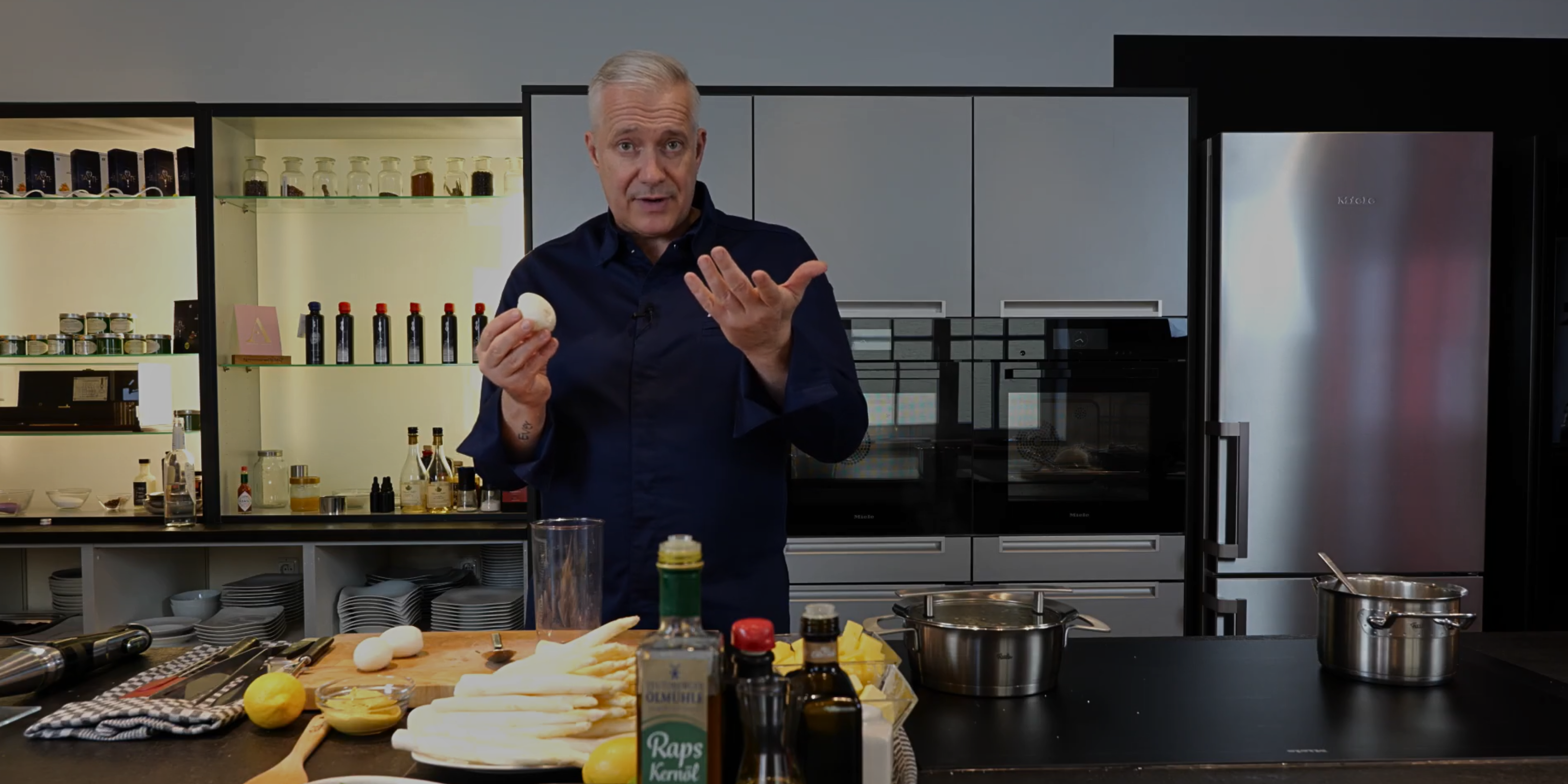 A man in a modern kitchen holds a peeled egg from Fissler cookware, with eggs, asparagus, and bottles on a wooden board in front of him.