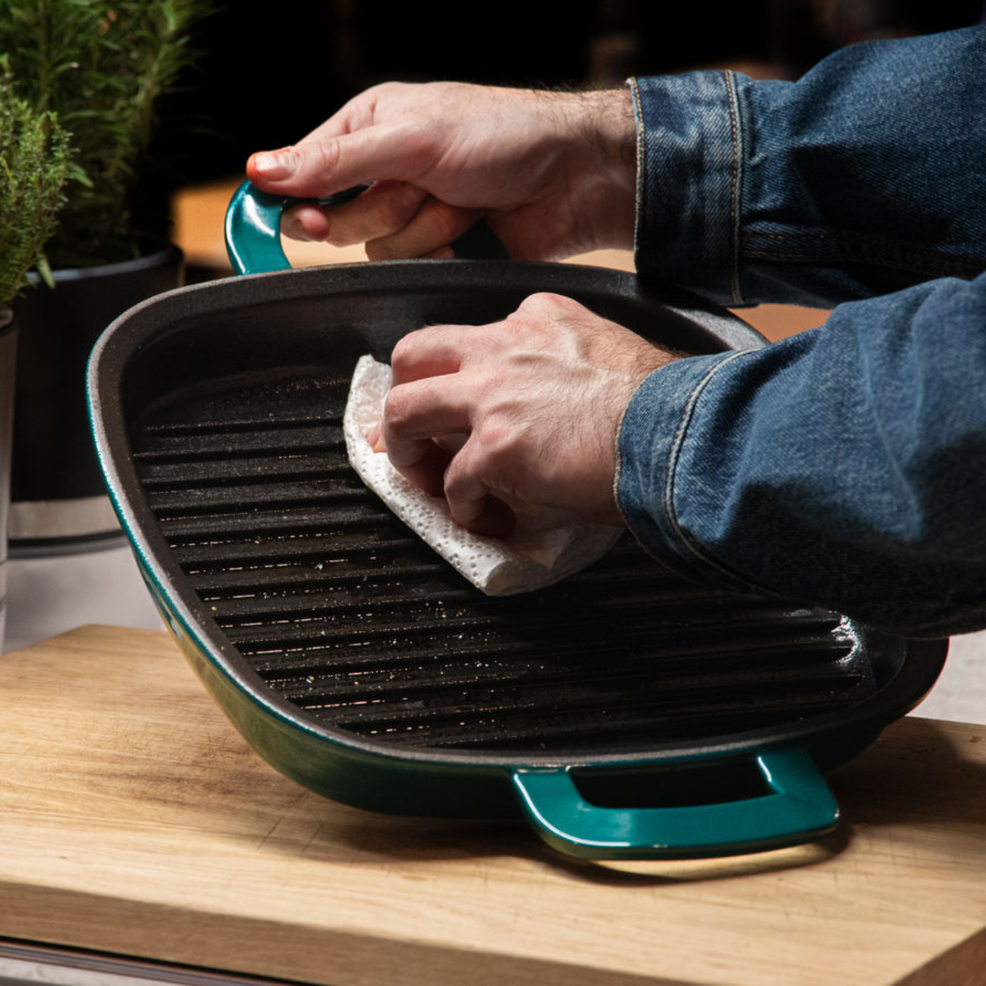 A person in a denim jacket cleans a high-quality cast iron grill pan on wood, with Fissler's quality and timeless German design nearby.