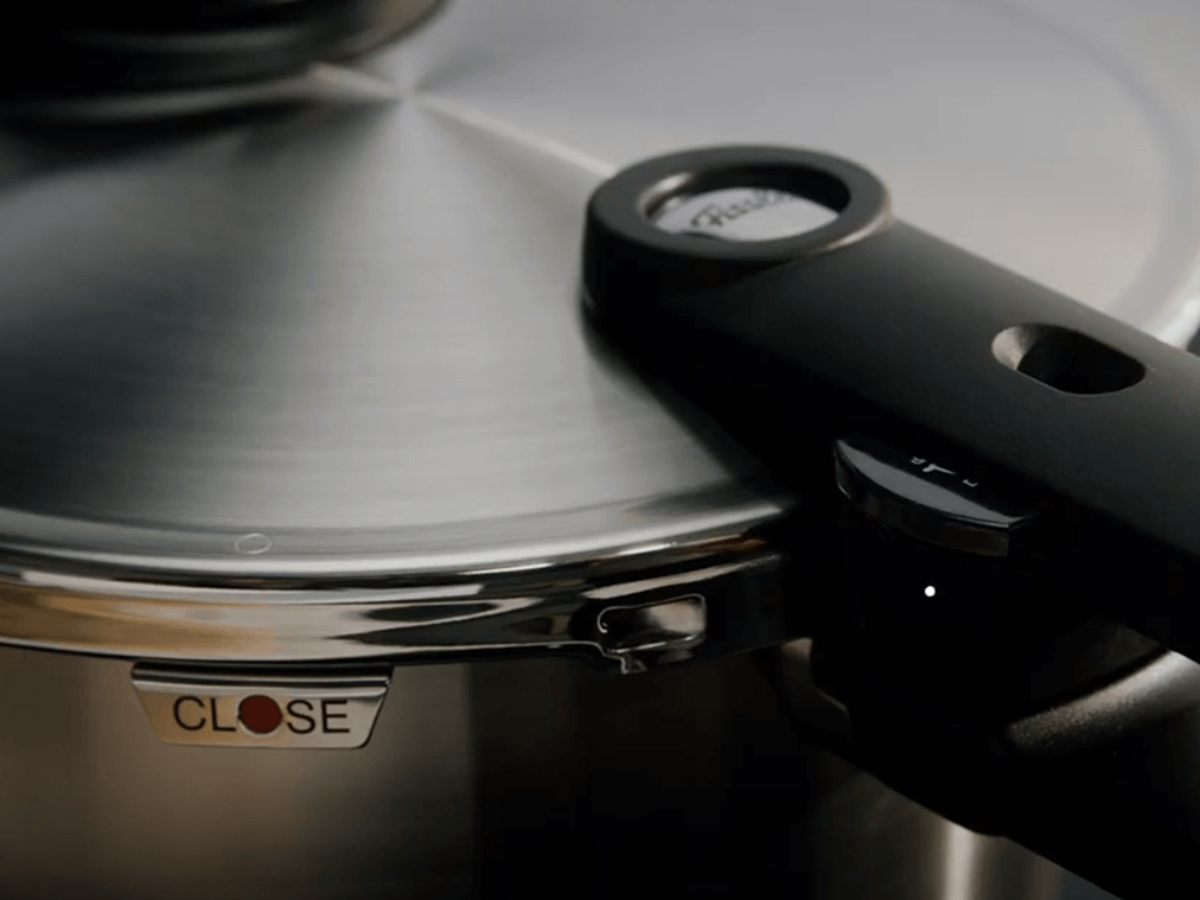 Nahaufnahme des Fissler Schnellkochtopfdeckels mit poliertem Edelstahl, ergonomischem Griff, Signaturknopf und CLOSE-Etikett in Rot und Schwarz.