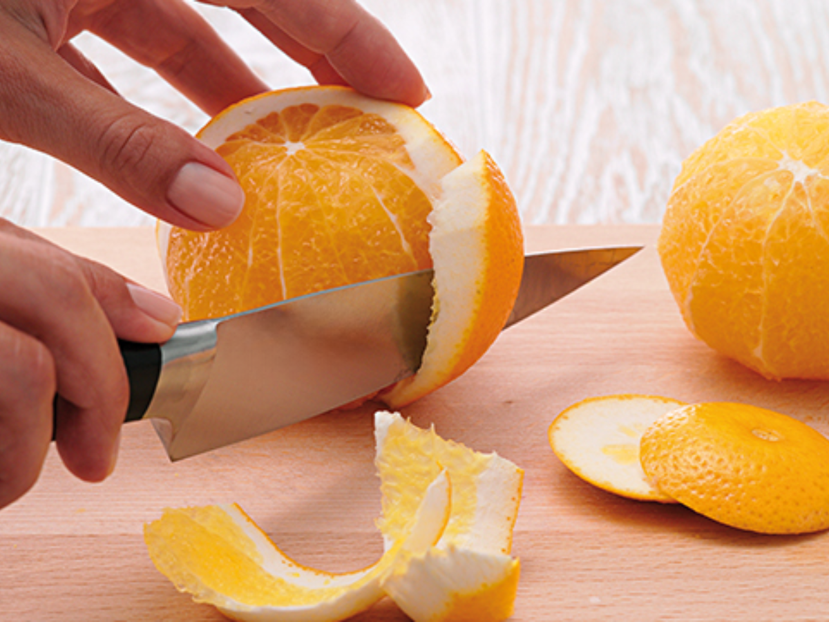 A person peels an orange with a Fissler kitchen knife on a wooden cutting board, with the peeled orange and peels lying next to it.