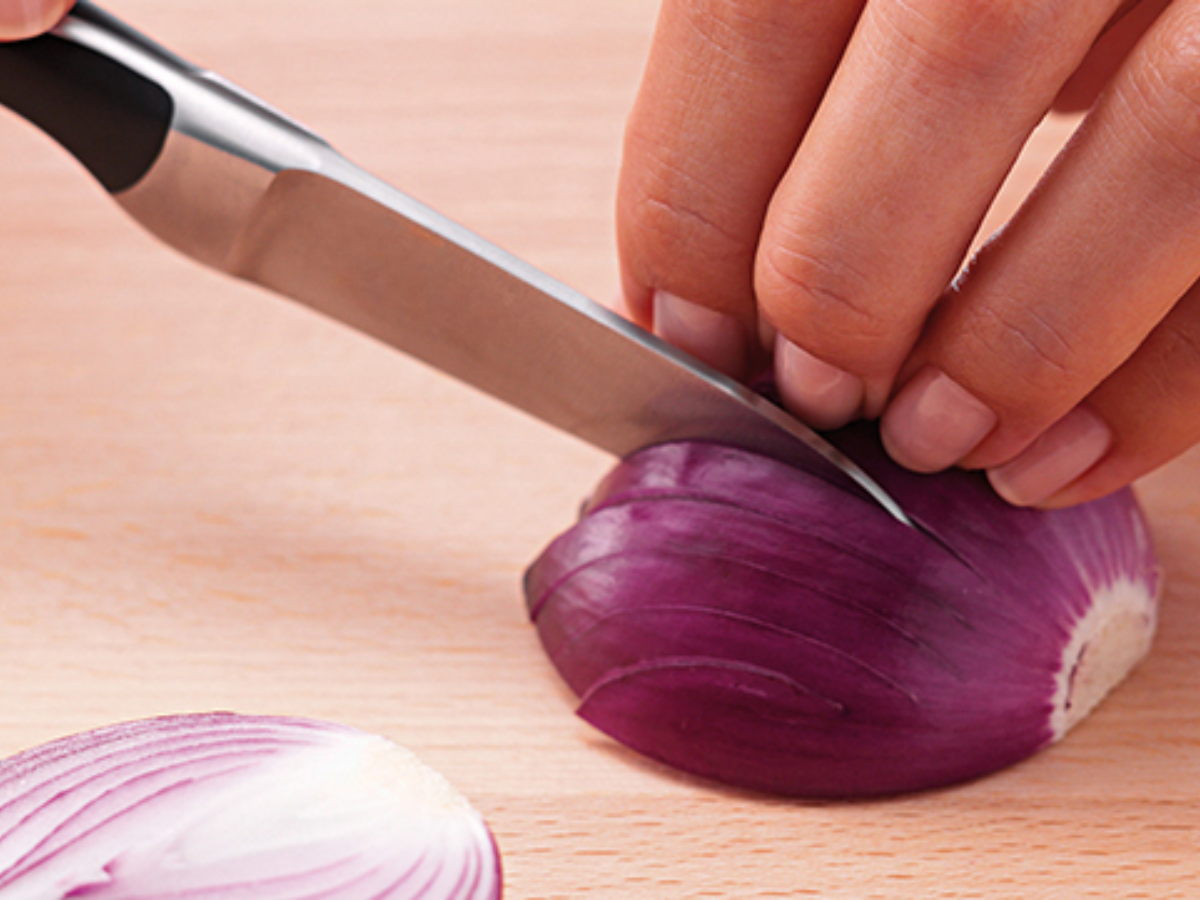 Nahaufnahme einer Hand, die fachmännisch eine rote Zwiebel mit einem Präzisions-Fissler-Messer auf einem hölzernen Schneidebrett schneidet.