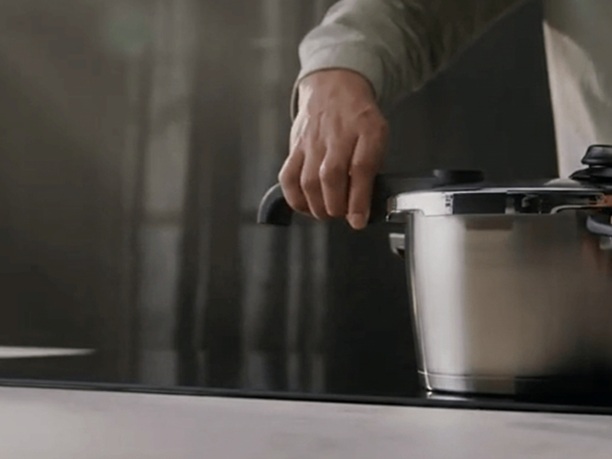 A person’s hand holds the ergonomic handle of a Fissler stainless steel pressure cooker, renowned for German precision engineering and timeless design, as it rests on a sleek dark kitchen counter.