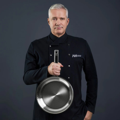 A distinguished chef in black holds a Fissler stainless steel frying pan, presenting German precision against a dark background.