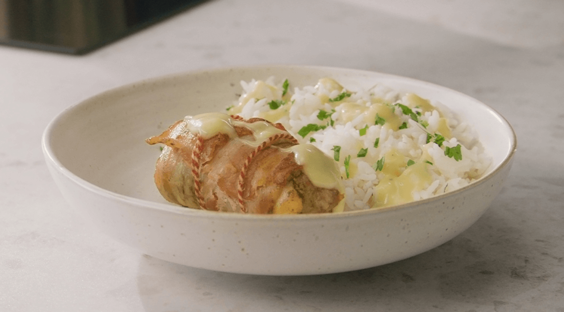 Weißer Reis mit Kräutern in einer Keramikschale neben mit Speck und Käse umwickeltem Hühnchen, serviert auf elegantem Fissler-Kochgeschirr.
