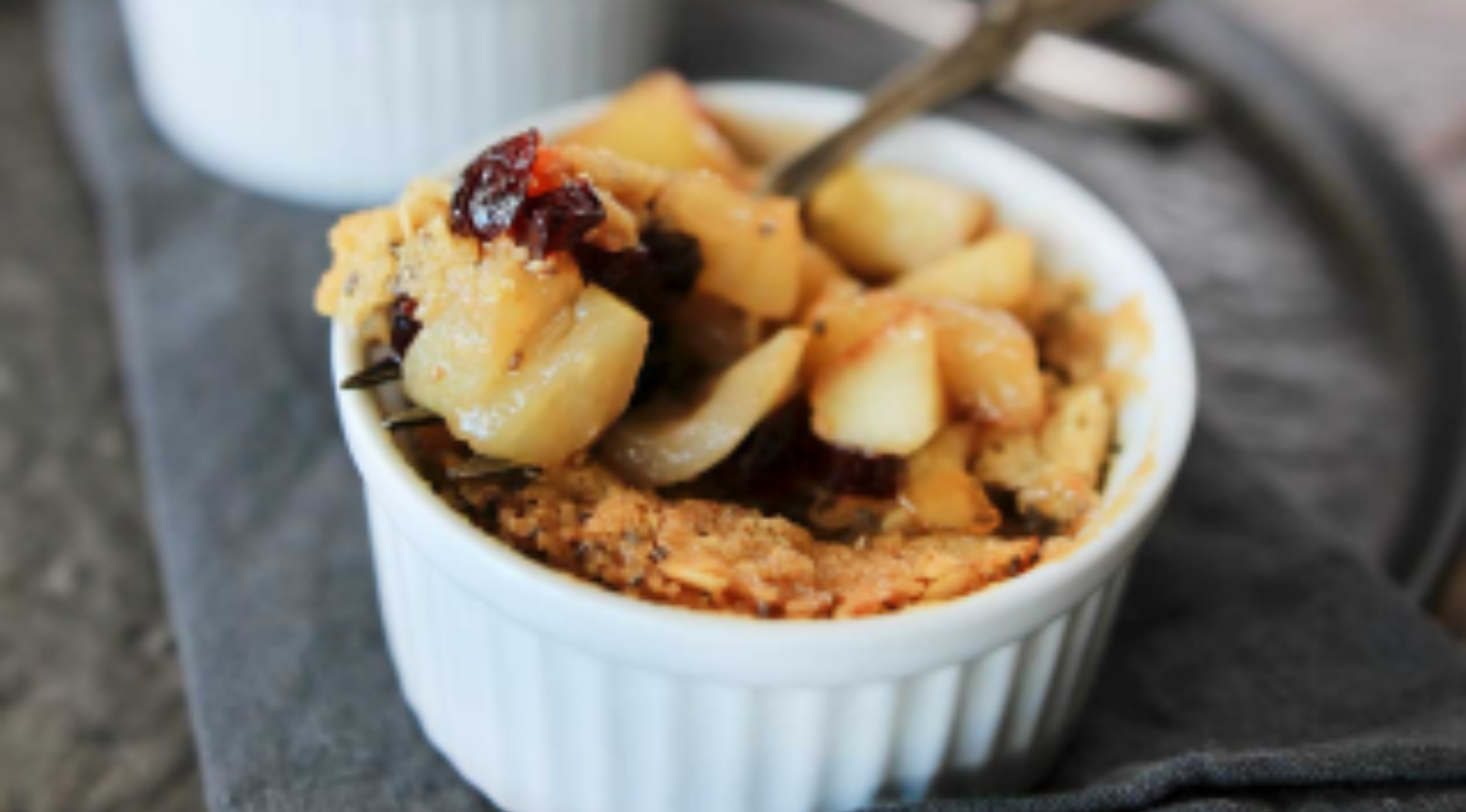 Apfelstreusel mit Äpfeln und Preiselbeeren in einer weißen Auflaufform, serviert mit einem Löffel auf einem grauen Tuch, das wie das Fissler-Kochgeschirr perfekt verarbeitet ist.