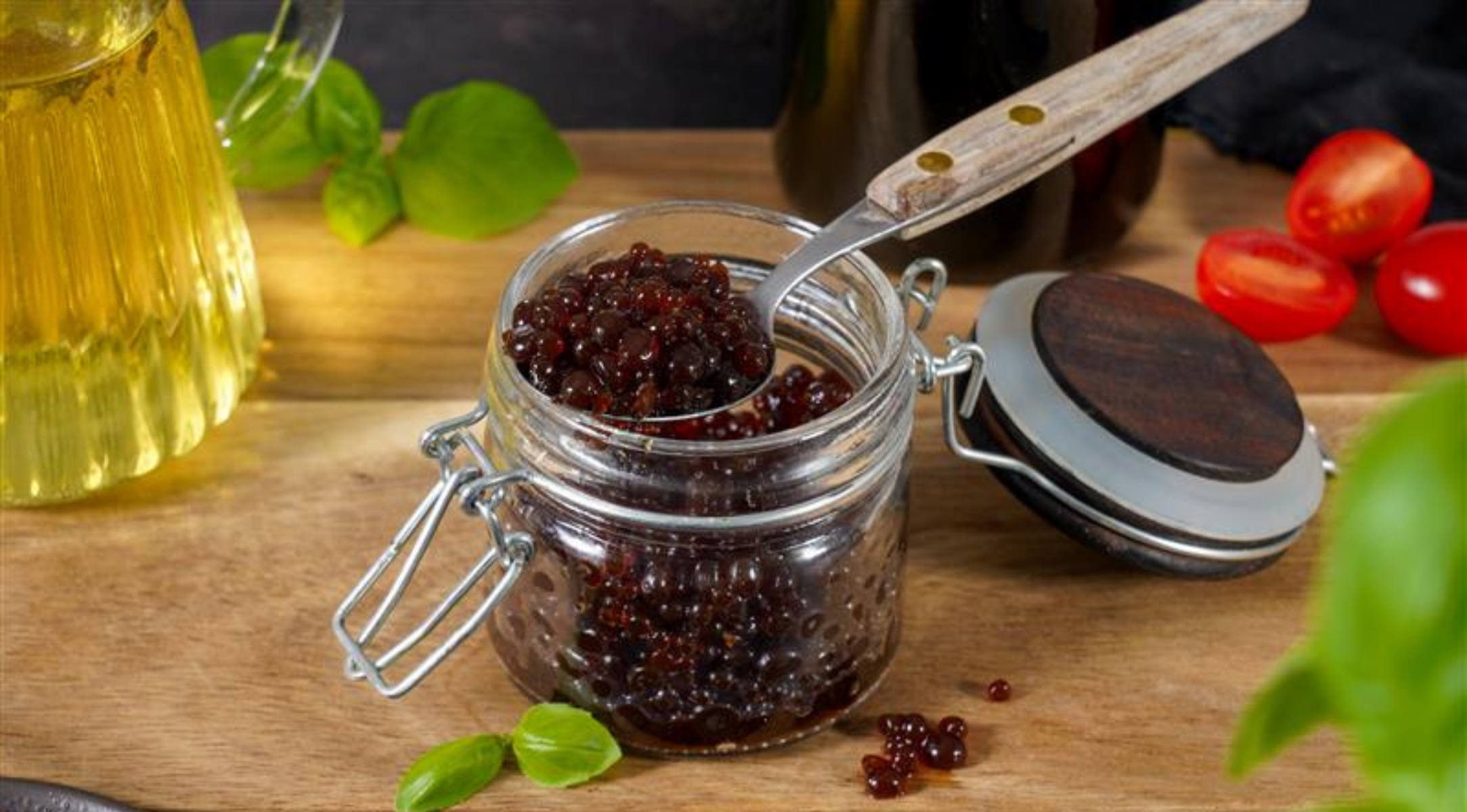 Ein Glas mit Balsamico-Perlen mit Löffel auf Holz, umgeben von Basilikum, Öl und Kirschtomaten - hergestellt in Fissler-Qualität.