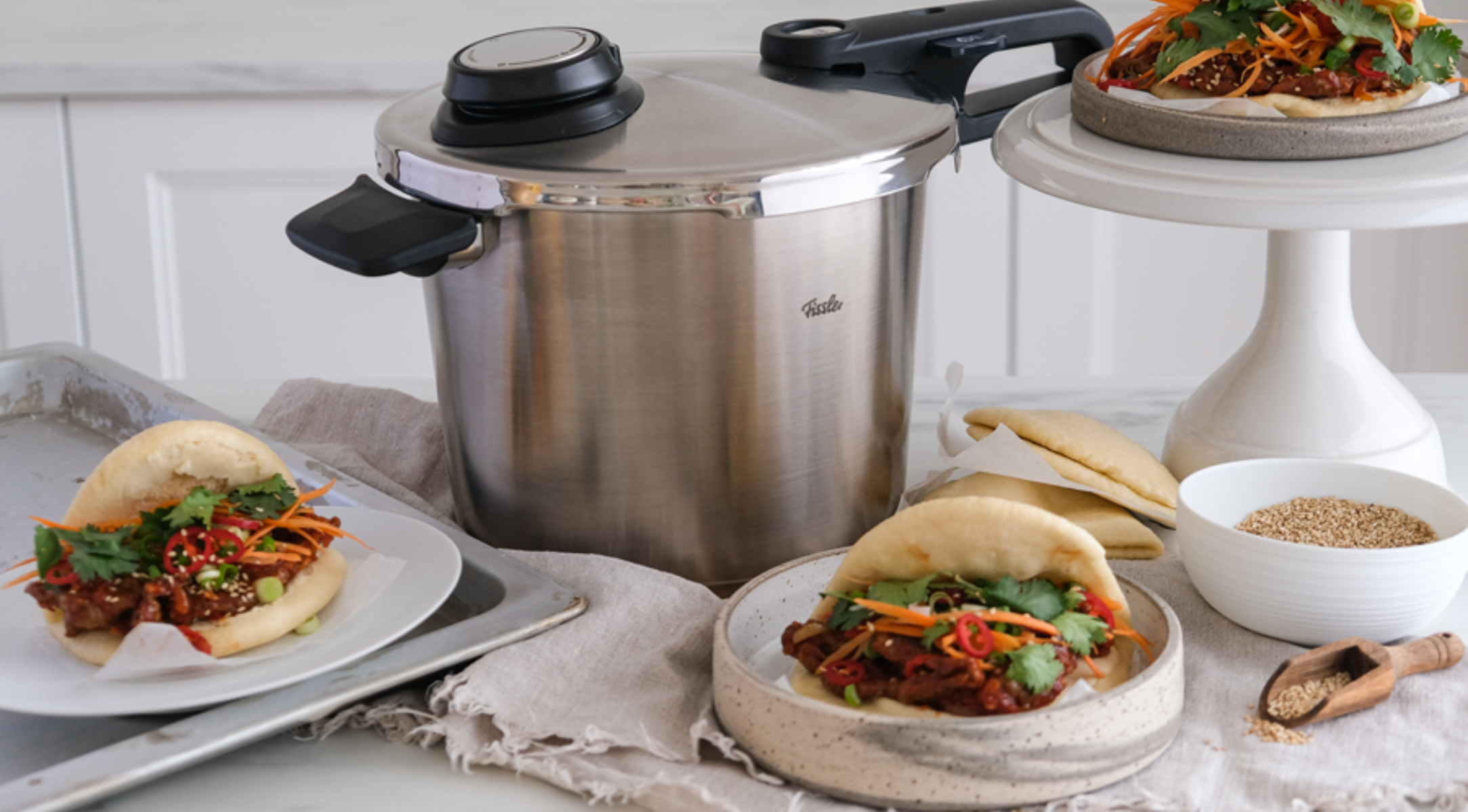 Fissler Schnellkochtopf aus Edelstahl auf einem Küchentisch mit Bao-Brötchen, frischen Füllungen, Sesamkörnern und einem Tablett mit weiteren Brötchen.