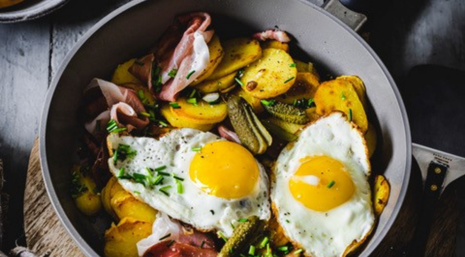 Zwei Eier, Bratkartoffeln, Schinken, Essiggurken und Schnittlauch in einer Fissler-Pfanne auf Holz - präzise gegart für zeitlosen Genuss.