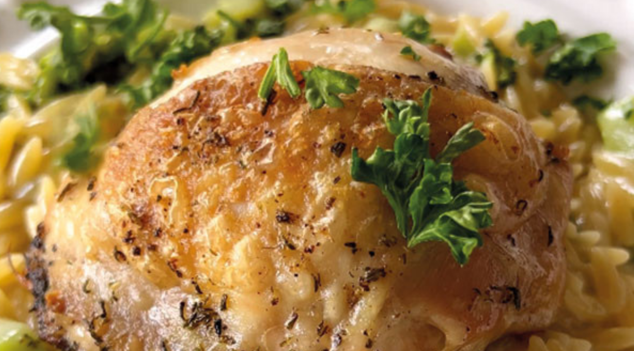 Ein perfekt gebratener Hähnchenschenkel mit Petersilie auf Kräuter-Orzo, elegant präsentiert auf einem weißen Teller - zubereitet im Fissler Kochgeschirr.