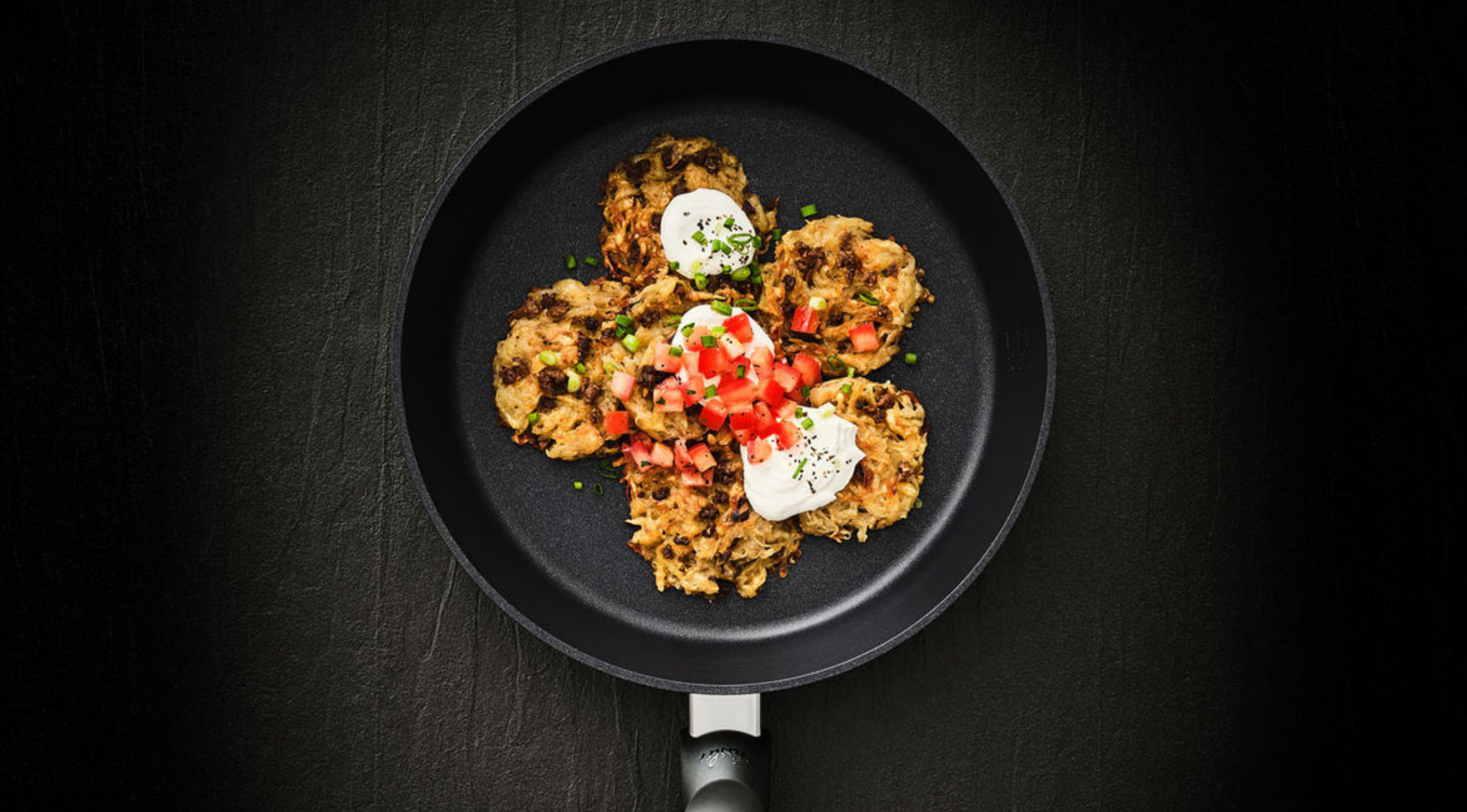 Eine Fissler Bratpfanne mit vier Gemüsepfannkuchen mit Sauerrahm, Tomaten und Schnittlauch auf einer raffinierten dunklen Oberfläche.