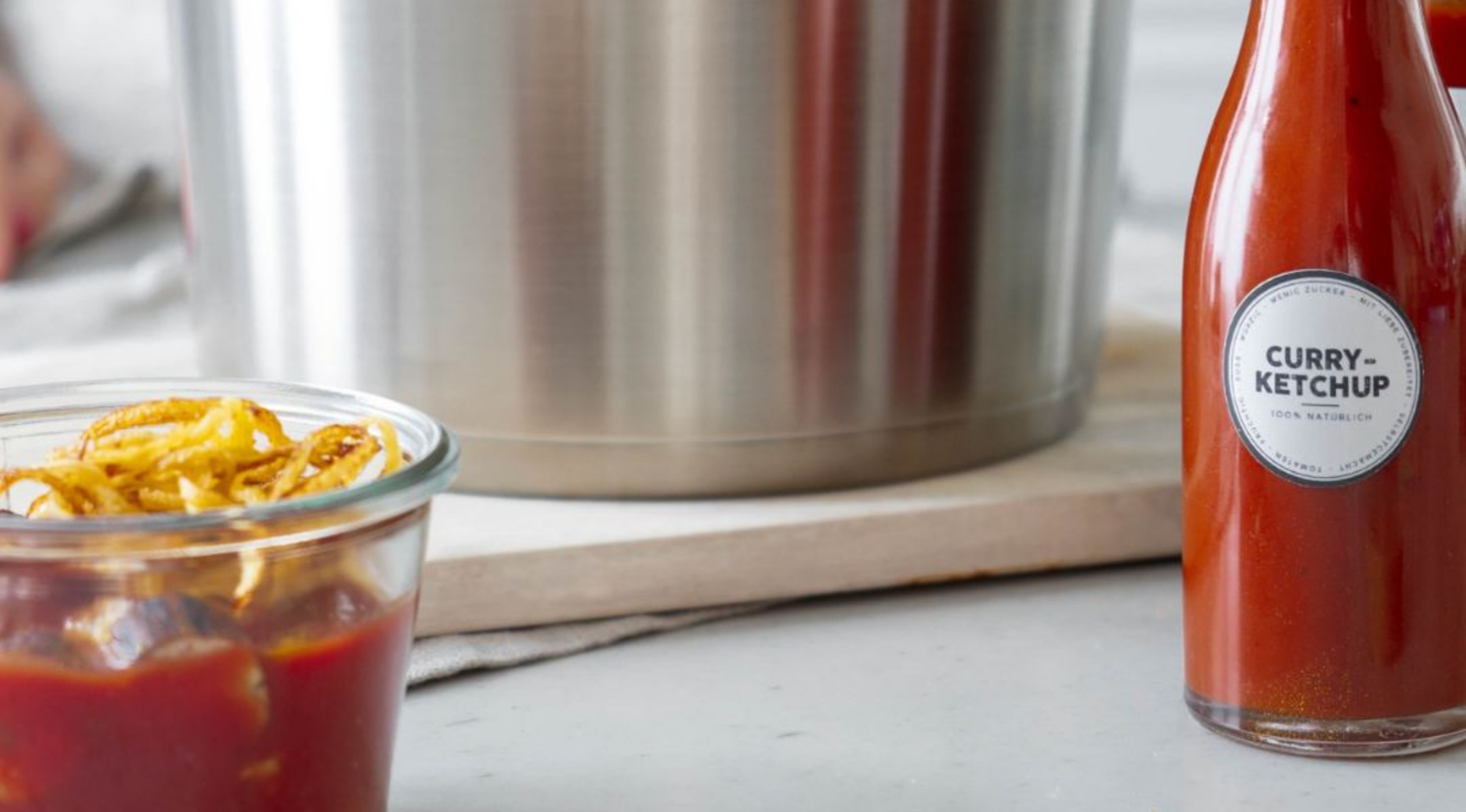 Ein Fissler-Edelstahltopf steht hinter einem Glas mit Pommes frites und Ketchup, einer Curry-Ketchup-Flasche und einem Holzbrett auf der Theke.