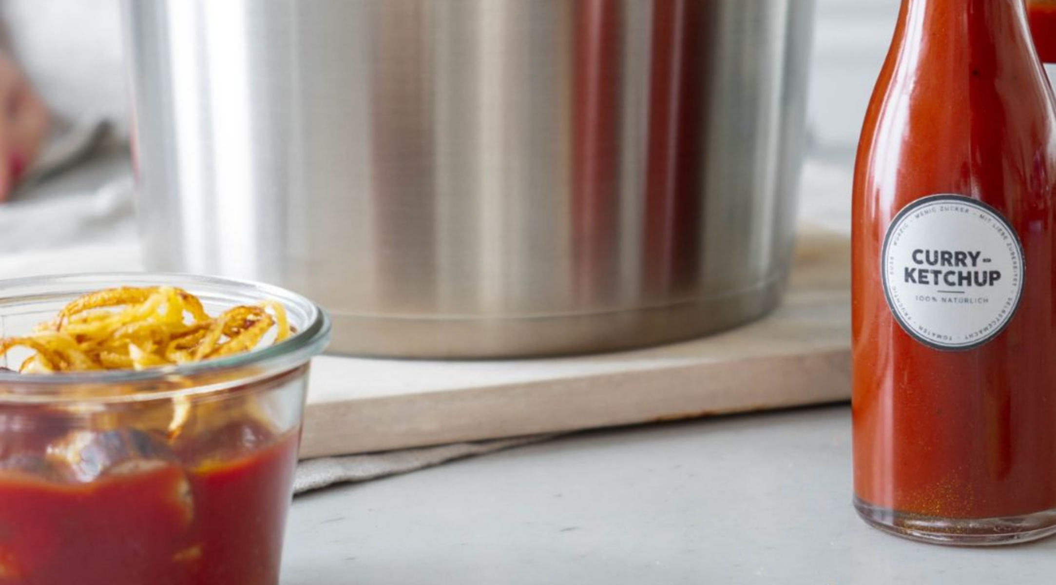 Ein Fissler-Edelstahltopf steht hinter einem Glas mit Pommes frites und Ketchup, einer Curry-Ketchup-Flasche und einem Holzbrett auf der Theke.