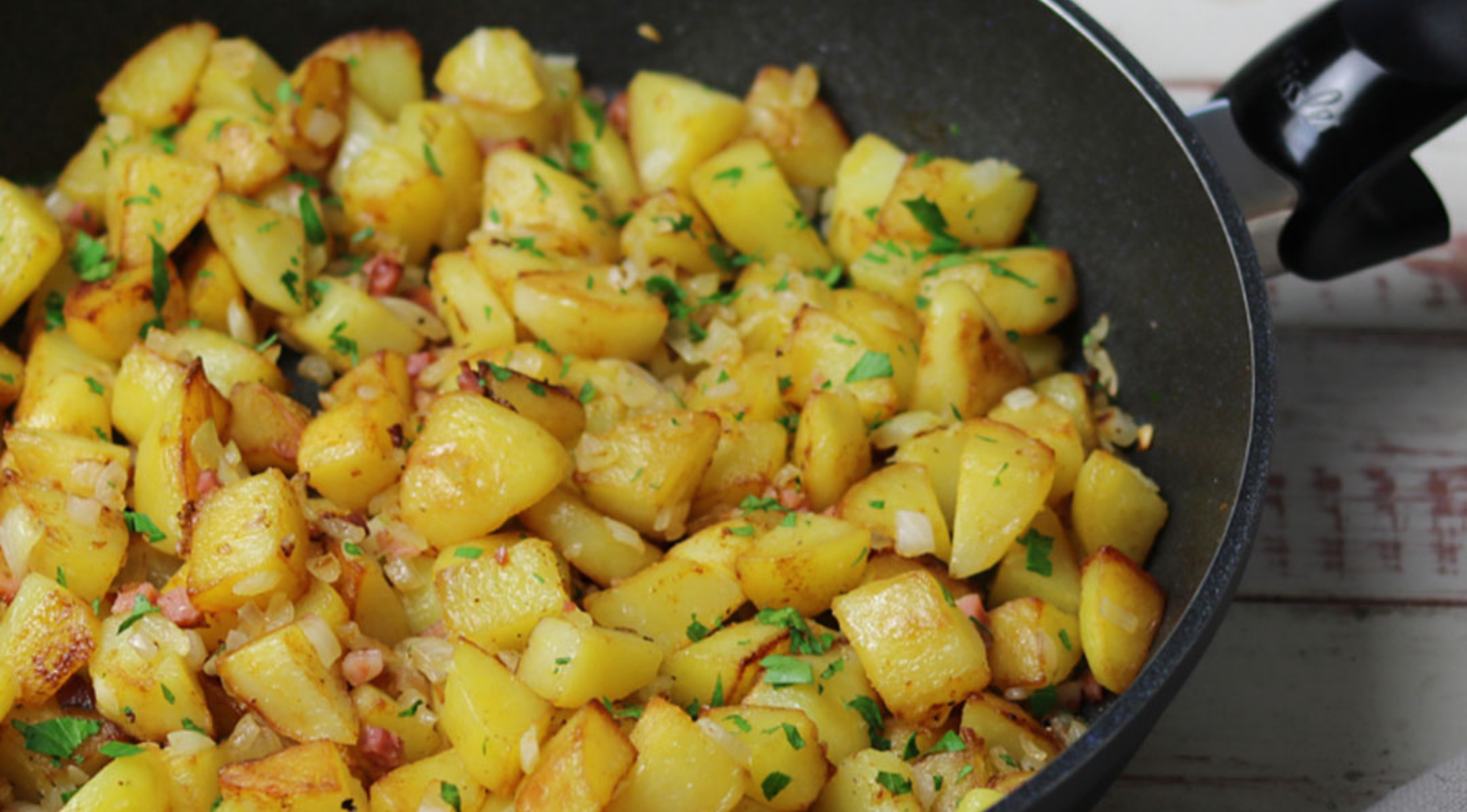 Knusprige, goldgelbe Kartoffelwürfel mit Kräutern und Zwiebeln in einer Fissler Präzisionspfanne auf einer hellen Holzoberfläche.