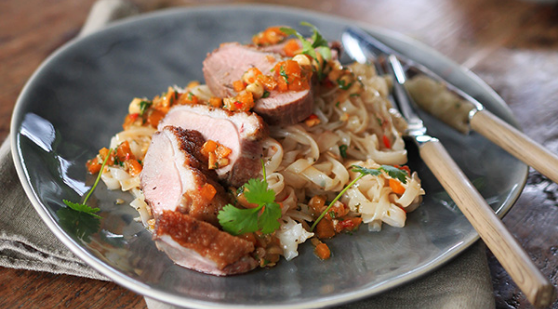 Gebratene Ente in Scheiben auf flachen Reisnudeln, frischen Kräutern und Gemüse auf einem grauen Teller, mit Fissler-Besteck auf Leinenserviette.