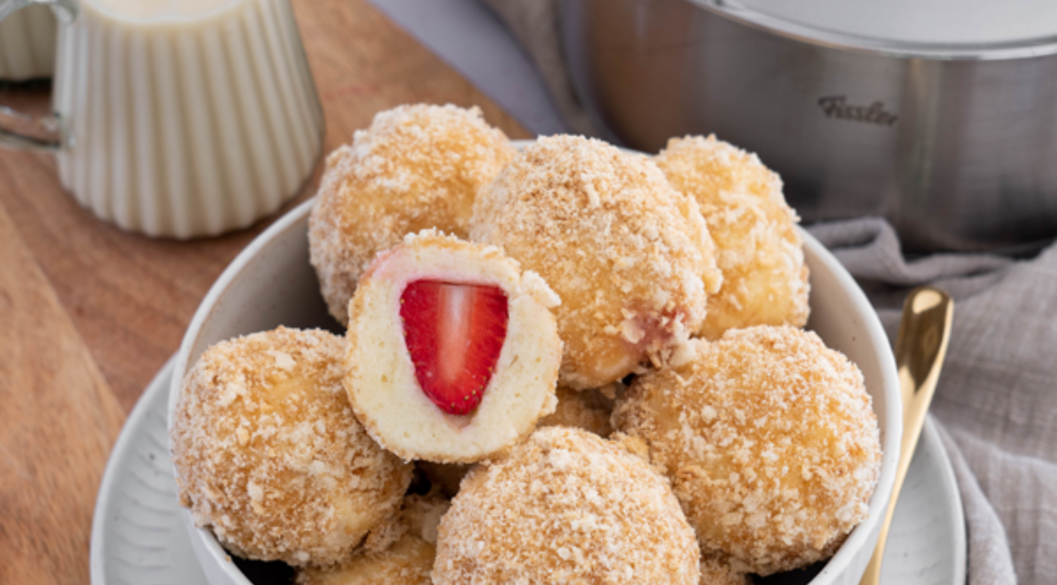 Mit Erdbeeren gefüllte Knödel in einer Schüssel, im Hintergrund ein Fissler-Edelstahltopf und ein Sahnekrug auf einer Holzfläche.