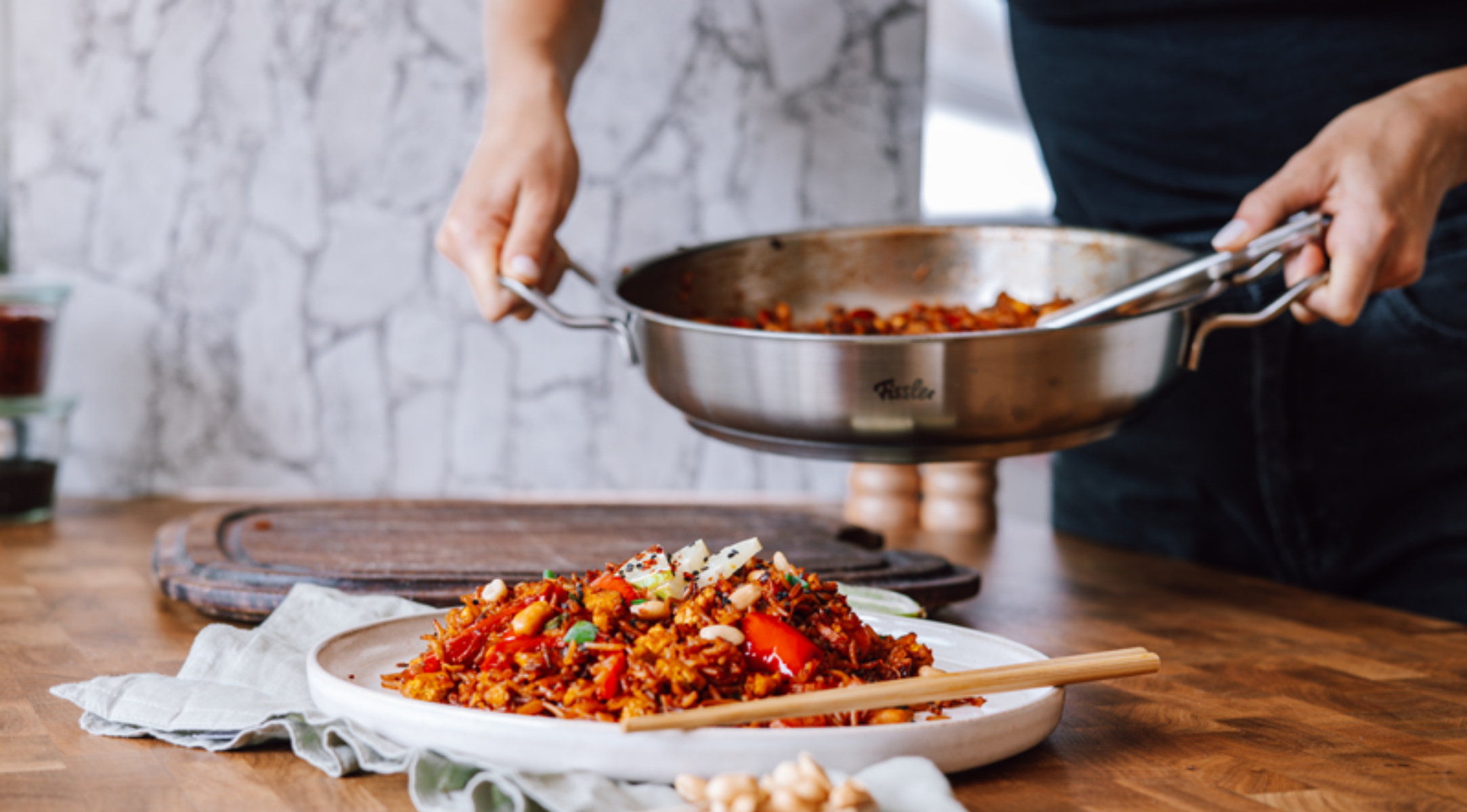 Eine Person serviert aus einer Fissler-Edelstahlpfanne pikante Pfannengerichte und Reis auf einen Teller, die Stäbchen liegen ordentlich auf dem Holztisch.