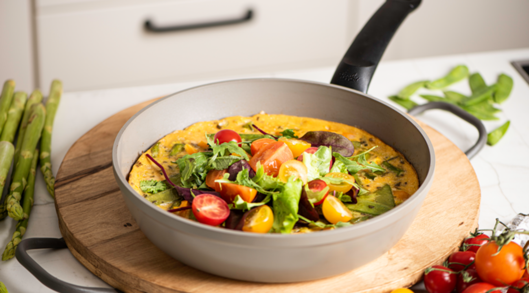 Eine lebendige Gemüse-Frittata in einer Fissler-Pfanne, belegt mit Gemüse und Tomaten, präsentiert auf Holz; daneben frische Produkte.