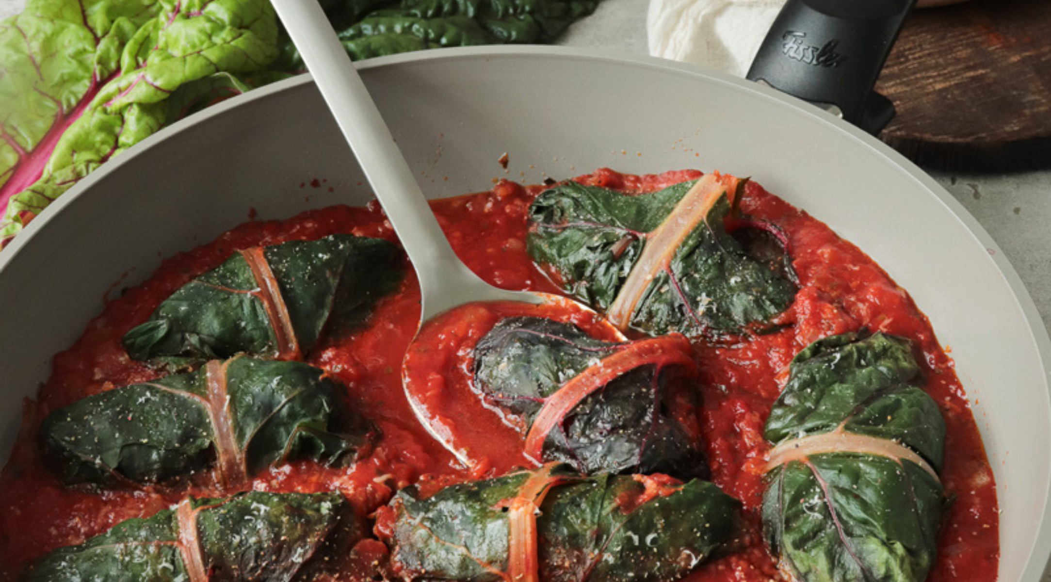 Fissler Edelstahlpfanne mit Mangoldröllchen in Tomatensauce; weißer Löffel und frische Mangoldblätter unterstreichen zeitloses deutsches Design.