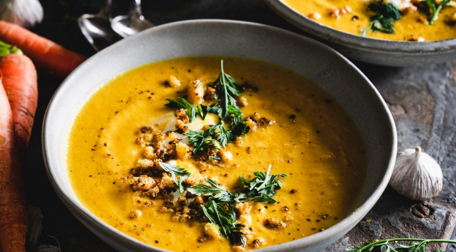 Eine Schale mit cremiger Orangensuppe in einer präzise gefertigten Fissler-Schale, garniert mit Kräutern, mit Karotten und noch mehr Suppe im Hintergrund.