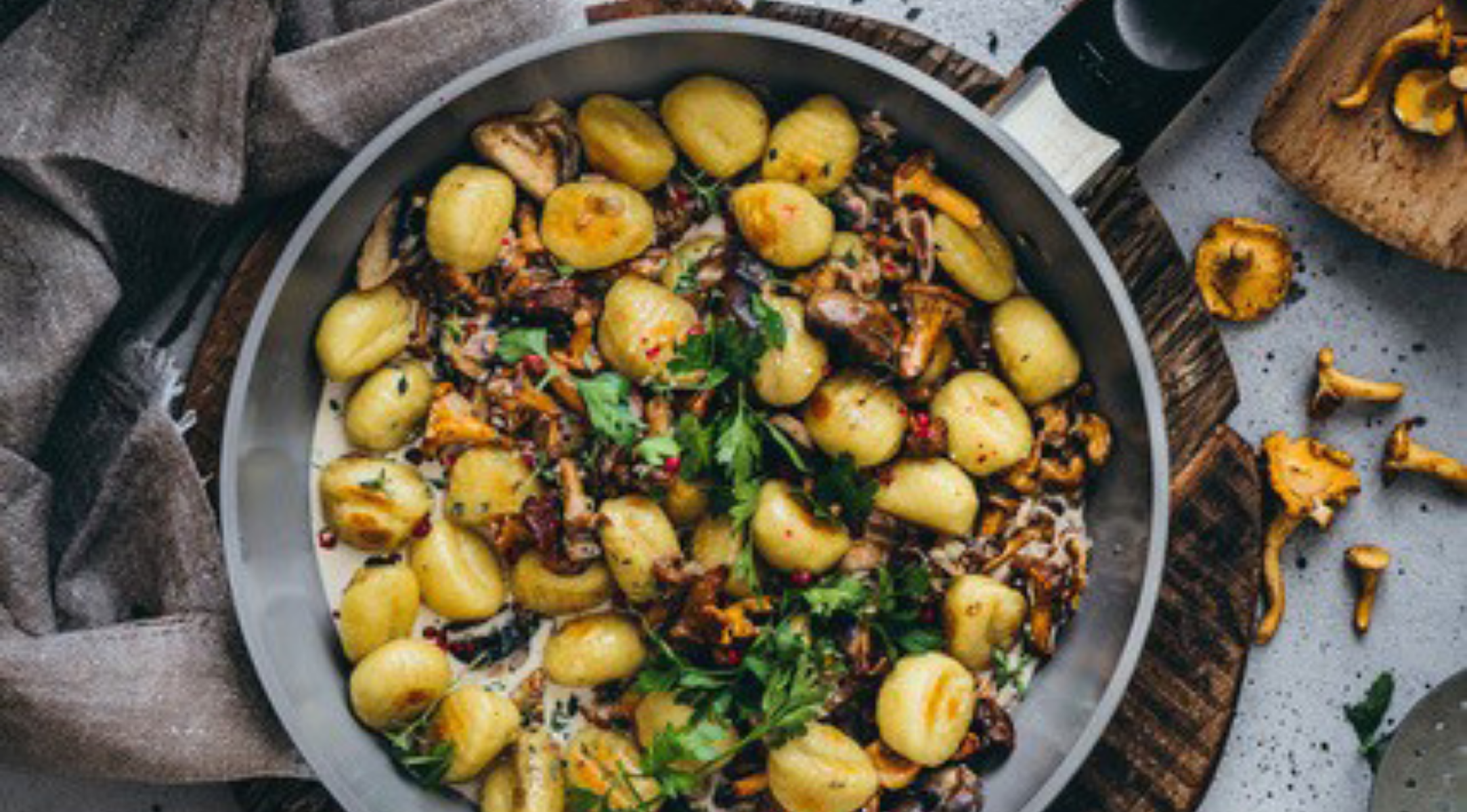 Fissler-Pfanne mit Gnocchi, Champignons, Sahnesauce, frischen Kräutern auf Holzbrett; daneben Stoffserviette und Champignons.