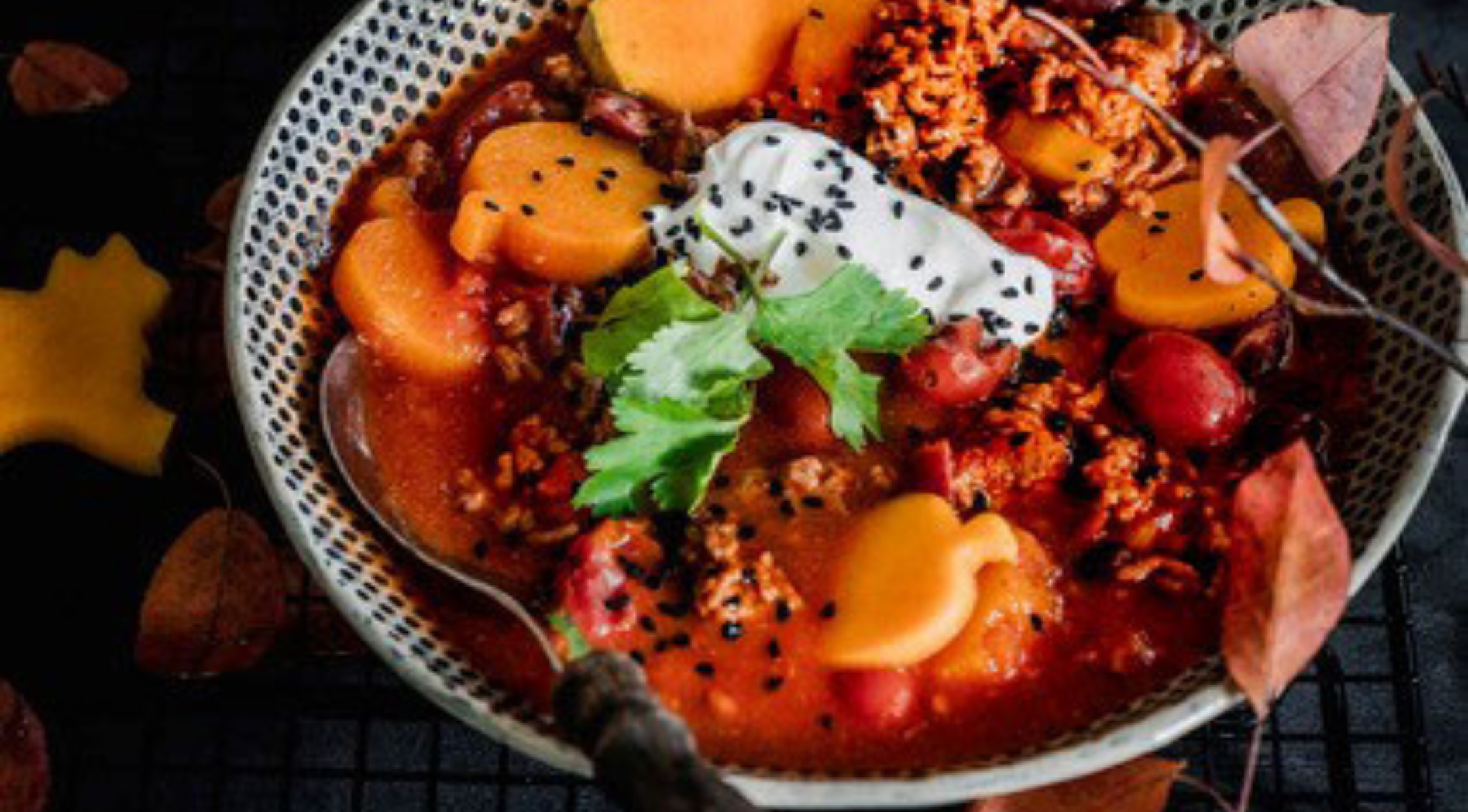 Chili in einer Fissler-Schale, garniert mit Sauerrahm und Kräutern, daneben ein hochwertiger Fissler-Löffel. Herbstblätter bilden eine gemütliche Kulisse.