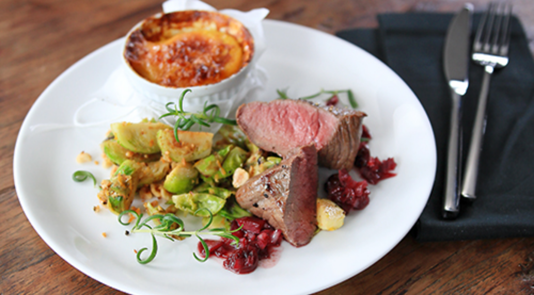 Ein weißer Teller mit gebratenem Rindfleisch, Gemüse, roter Sauce und Gratin steht auf Holz. Rechts ein rostfreies Besteck und eine Serviette von Fissler.