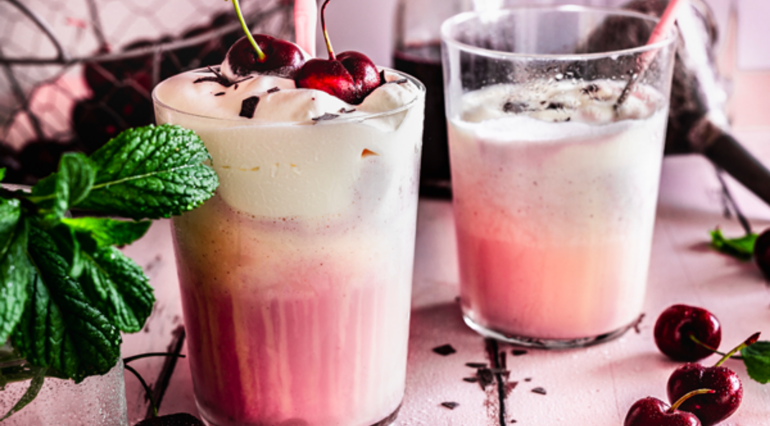 Zwei Milchshakes in eleganten Gläsern mit Schlagsahne und Kirschen, daneben Minze und Schokolade, auf einem raffinierten rosa Hintergrund - ganz im Stil von Fissler.