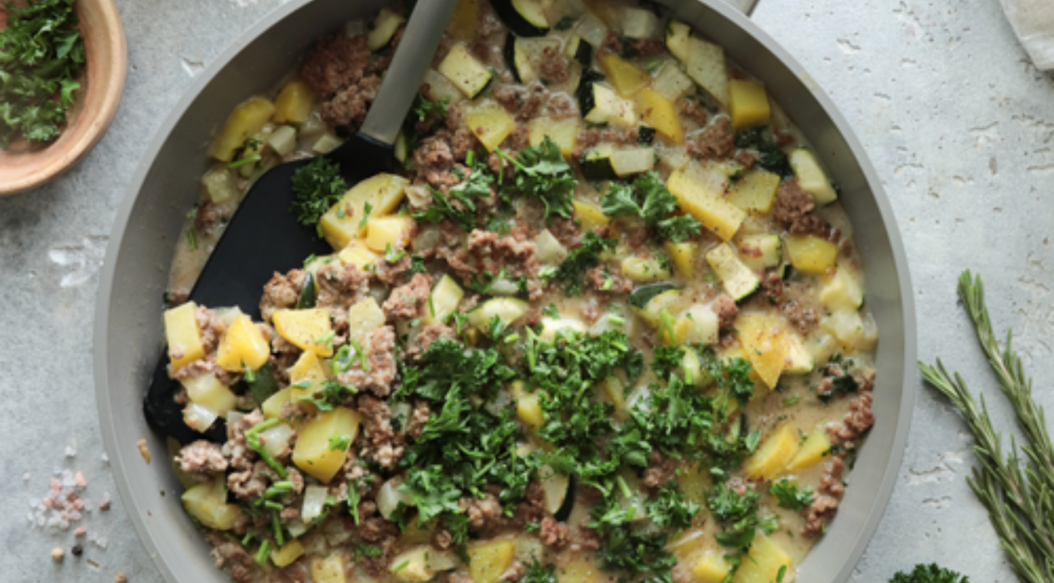 Eine Fissler-Pfanne mit Hackfleisch, Kartoffeln und Kräutern, garniert mit Petersilie. Servierbesteck liegt drinnen, frische Kräuter daneben.