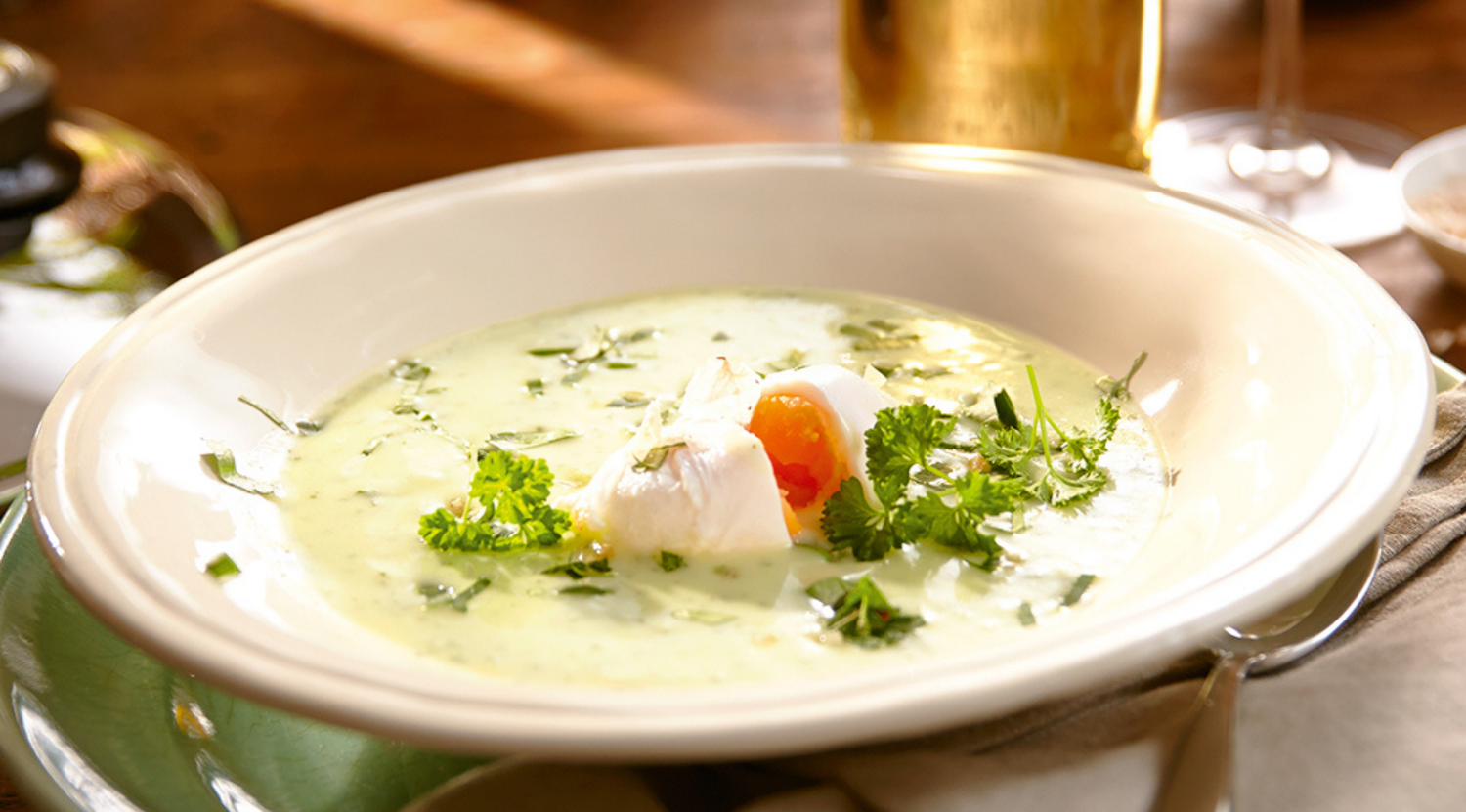 Grüne Cremesuppe, garniert mit Kräutern und einem pochierten Ei in einer weißen Schale, präsentiert auf einem eleganten Tisch mit Fissler-Kochgeschirr.